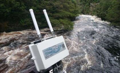 The UQ-developed flood tool monitoring the Gordon River, Tasmania. A white camera-looking object with two antennas sits above fast flowing brown water with bushland in the background.